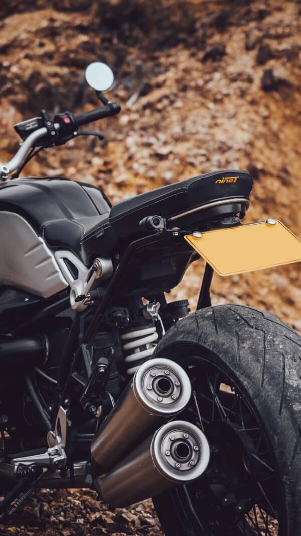 A close-up image of a part of a motorcycle, showcasing the rear wheel, twin exhaust pipes, and a portion of the seat with the logo "nineT" embossed on it, against a blurred background of autumn leaves.